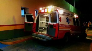 Una ambulancia de Protección Civil y Bomberos con las puertas traseras abiertas y luces encendidas está estacionada de noche frente a un edificio.