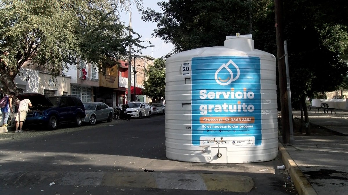 Primer plano de una llave de paso metálica conectada a un gran tinaco blanco en una calle residencial, con autos y casas de fondo desenfocados.