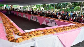 Una larguísima Rosca de Reyes se extiende sobre mesas alineadas en una plaza pública, rodeada por una multitud de personas que esperan su turno.