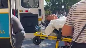 Paramédicos trasladan a un hombre en una camilla amarilla hacia una ambulancia abierta; el paciente lleva una máscara de oxígeno y está consciente