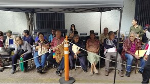 Un grupo de adultos mayores espera sentado bajo una carpa gris en una zona delimitada por cuerdas, algunos sostienen carpetas con documentos.