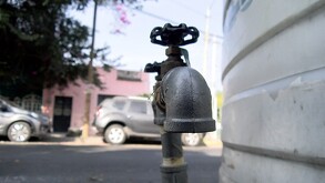 Primer plano de una llave de paso metálica conectada a un gran tinaco blanco en una calle residencial, con autos y casas de fondo desenfocados.