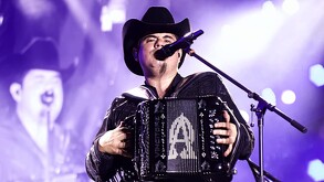 El cantante Alfredo Olivas, con sombrero negro y traje bordado, toca su acordeón frente a un micrófono bajo las intensas luces de un escenario.