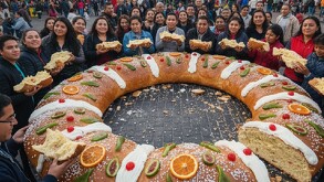 Se ve una rosca de Reyes gigante y mucha gente alrededor con un pedazo de pan en la mano.