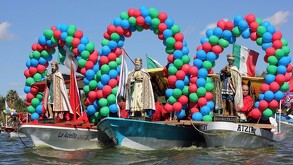 Tres lanchas decoradas con grandes arcos de globos de colores transportan estatuas de los Reyes Magos sobre el agua durante un desfile festivo.