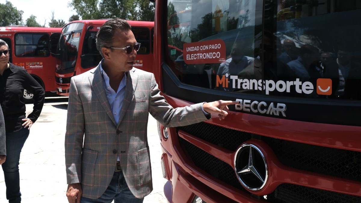 Pablo Lemus, con saco gris a cuadros y lentes de sol, señala el frente de una unidad roja de 