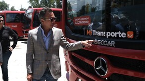 Pablo Lemus, con saco gris a cuadros y lentes de sol, señala el frente de una unidad roja de 