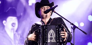 El cantante Alfredo Olivas, con sombrero negro y traje bordado, toca su acordeón frente a un micrófono bajo las intensas luces de un escenario.
