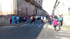 Una larga fila de personas espera sobre una acera frente a un edificio antiguo de cantera rosa en una calle peatonal bajo la luz del sol.