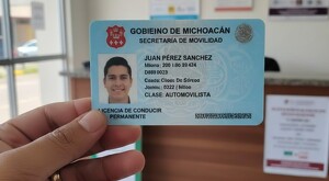 Persona sosteniendo una licencia de conducir con datos y la foto de un hombre viendo de frente con una oficina de gobierno de fondo