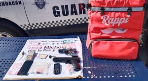 Mochila de reparto de Rappi junto a artefactos asegurados por la Policía de Michoacán delante de una patrulla