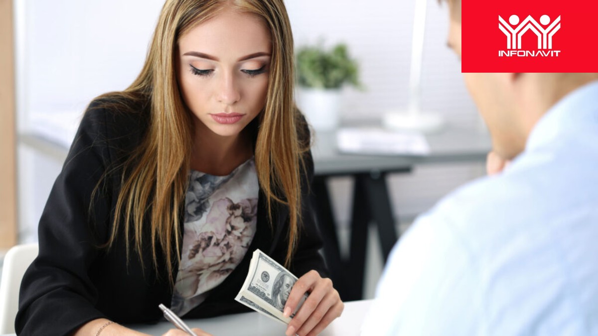 Una mujer joven con chaqueta negra sostiene un fajo de billetes de cien dólares mientras firma un documento; el logo rojo de Infonavit está arriba