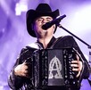 El cantante Alfredo Olivas, con sombrero negro y traje bordado, toca su acordeón frente a un micrófono bajo las intensas luces de un escenario.