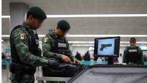 Dos militares con uniformes pixelados y boinas verdes inspeccionan una maleta en un aeropuerto; en una pantalla cercana se muestra la silueta de una pistola.