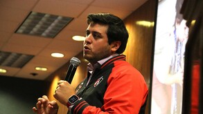 Un hombre joven con una chaqueta deportiva del equipo Atlas FC habla por un micrófono durante una conferencia en una sala con paneles de madera.