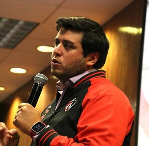 Un hombre joven con una chaqueta deportiva del equipo Atlas FC habla por un micrófono durante una conferencia en una sala con paneles de madera.