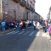 Una larga fila de personas espera sobre una acera frente a un edificio antiguo de cantera rosa en una calle peatonal bajo la luz del sol.