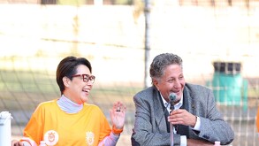 Juan José Frangie, con saco gris, y una mujer con gafas y camiseta naranja sonríen alegremente durante un evento público en una cancha deportiva.