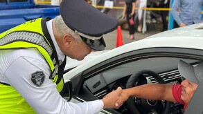 Un oficial de la Policía Vial, con chaleco fluorescente, estrecha la mano de un conductor que sonríe desde el interior de su vehículo blanco.