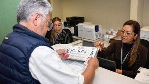 En una oficina gubernamental, una funcionaria entrega un juego de placas vehiculares nuevas a un ciudadano mayor que las recibe sobre el mostrador.