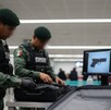 Dos militares con uniformes pixelados y boinas verdes inspeccionan una maleta en un aeropuerto; en una pantalla cercana se muestra la silueta de una pistola.