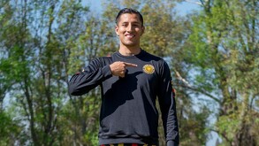 El futbolista Carlos Cisneros sonríe al aire libre mientras señala con su dedo índice el escudo de los Leones Negros en su sudadera negra.