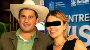 Un hombre con sombrero de palma y camisa verde posa sonriente junto a una mujer con gafas oscuras, frente a un banner de la Secretaría del Migrante.