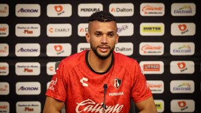 El futbolista Matheus Dória, con la camiseta roja de entrenamiento del Atlas FC, habla frente a un muro de patrocinadores durante una rueda de prensa.