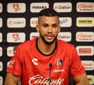El futbolista Matheus Dória, con la camiseta roja de entrenamiento del Atlas FC, habla frente a un muro de patrocinadores durante una rueda de prensa.