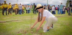 Una mujer con sombrero y ropa clara planta un pequeño árbol en un campo, mientras al fondo un grupo de brigadistas con cascos amarillos observa.