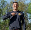 El futbolista Carlos Cisneros sonríe al aire libre mientras señala con su dedo índice el escudo de los Leones Negros en su sudadera negra.