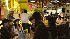Hombres y mujeres conviviendo al interior de un bar decorado con temas mexicanos, una banda tocando al fondo y bebidas alcohólicas en las mesas
