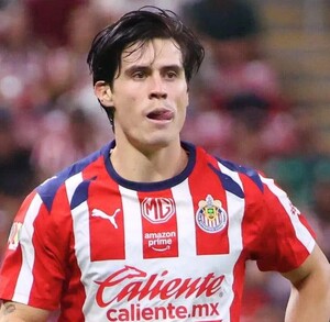 El futbolista Ricardo Campillo, con el uniforme de local de las Chivas, aparece en el campo de juego con una expresión de esfuerzo y la lengua fuera.