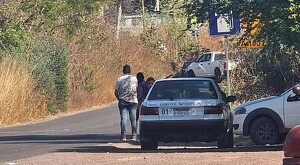 Taxi da la marca Nissan tipo Tsuru estacionado en un crucero con letreros viales, personas de pie y otros vehículos alrededor de matorrales