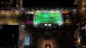 Vista aérea nocturna de una cancha de fútbol rápido iluminada en una plaza pública de Guadalajara, rodeada de edificios y personas observando.