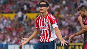 Un jugador de las Chivas con uniforme rojiblanco y gafete de capitán abre los brazos en el campo; sus ojos están cubiertos por una barra negra.