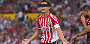 Un jugador de las Chivas con uniforme rojiblanco y gafete de capitán abre los brazos en el campo; sus ojos están cubiertos por una barra negra.