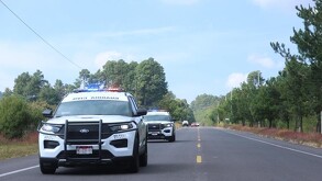 Convoy de la Guardia Civil patrulla un camino rural de tierra; agentes a pie y en camionetas avanzan entre maleza seca bajo un cielo despejado.