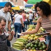 Mujer con ropa provocativa ofreciendo un boleto de rifa colombiana a un vendedor de un mercado con frutas, verduras y personas alrededor