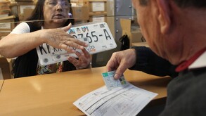 Una empleada tras una ventanilla entrega un juego de placas vehiculares nuevas a un ciudadano que sostiene una tarjeta de circulación y un recibo.