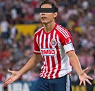 Un jugador de las Chivas con uniforme rojiblanco y gafete de capitán abre los brazos en el campo; sus ojos están cubiertos por una barra negra.