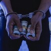 Primer plano de las manos de una persona esposada que sostiene varios rollos de billetes de dólares estadounidenses, bajo una iluminación fría.