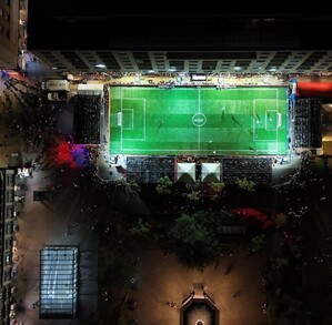 Vista aérea nocturna de una cancha de fútbol rápido iluminada en una plaza pública de Guadalajara, rodeada de edificios y personas observando.