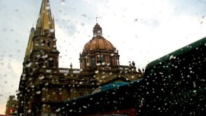 Vista desenfocada de la Catedral de Guadalajara a través de un cristal cubierto de gotas de lluvia, con la silueta de un autobús en primer plano.