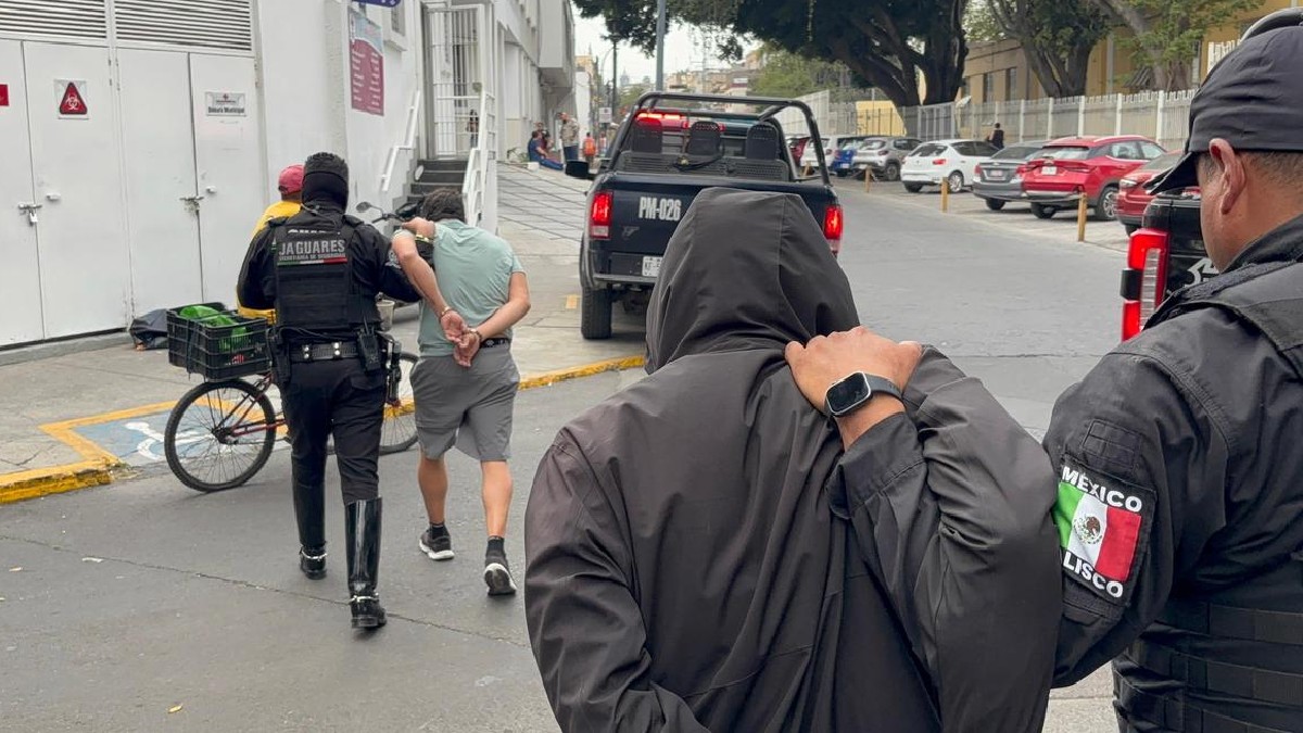 Hombres de espaldas siendo arrestados y llevados por policías de Jalisco con una patrulla estacionada y personas alrededor