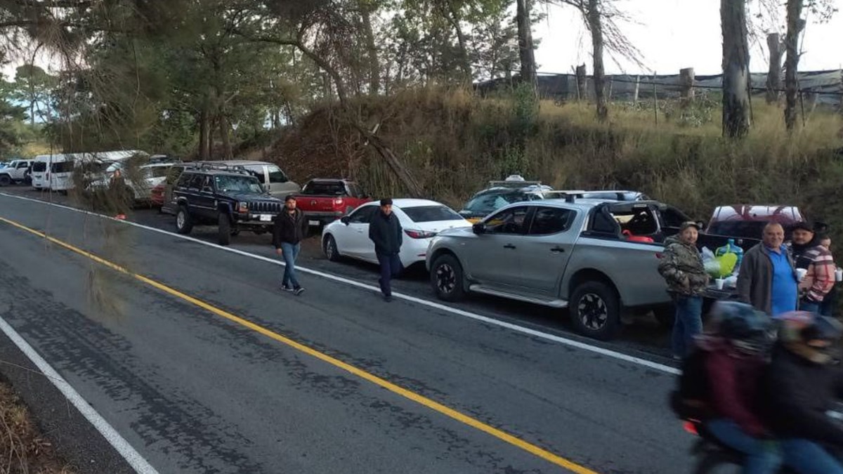 Fila de automóviles detenidos en una carretera con matorrales y personas alrededor. Una moto pasando en la esquina derecha