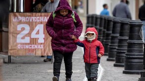 Una mujer sosteniendo de la mano a un niño mientras caminan juntos por la calle usando abrigos. Un letrero de promoción de fondo