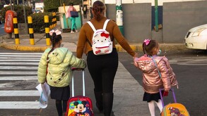 Se observa a una mujer que lleva de la mano a dos niñas que traen mochilas.