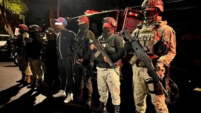 Un hombre con los ojos cubiertos por una barra blanca es custodiado por agentes fuertemente armados del Ejército y la Guardia Nacional de noche.