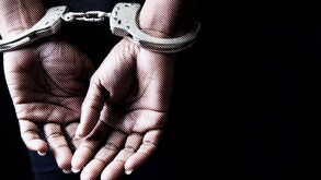 Primer plano detallado de las manos de una persona hacia atrás y sujetas con esposas metálicas, sobre un fondo negro sólido.